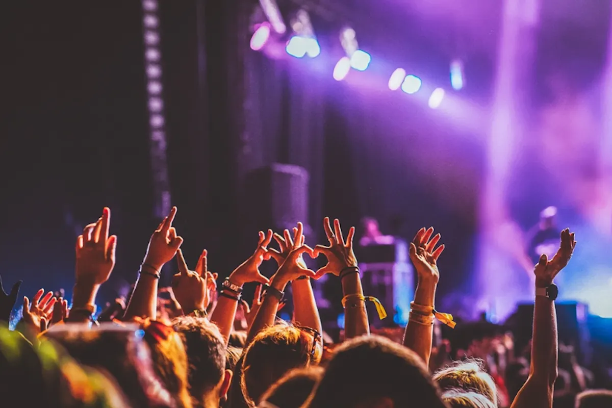 Live Event Audience at a concert with colorful lights in the background. People raise their hands, some forming a heart shape. The atmosphere is energetic and lively.