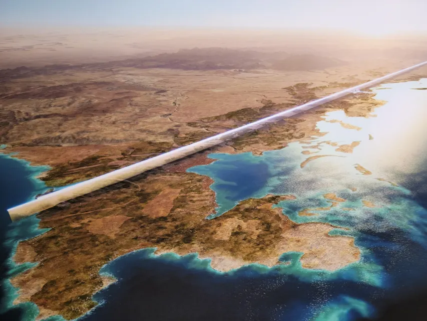 Aerial view of a long, futuristic linear structure cutting across an expansive desert landscape, bordered by the sea. The structure is illuminated by sunlight, highlighting its sleek design against the natural terrain.