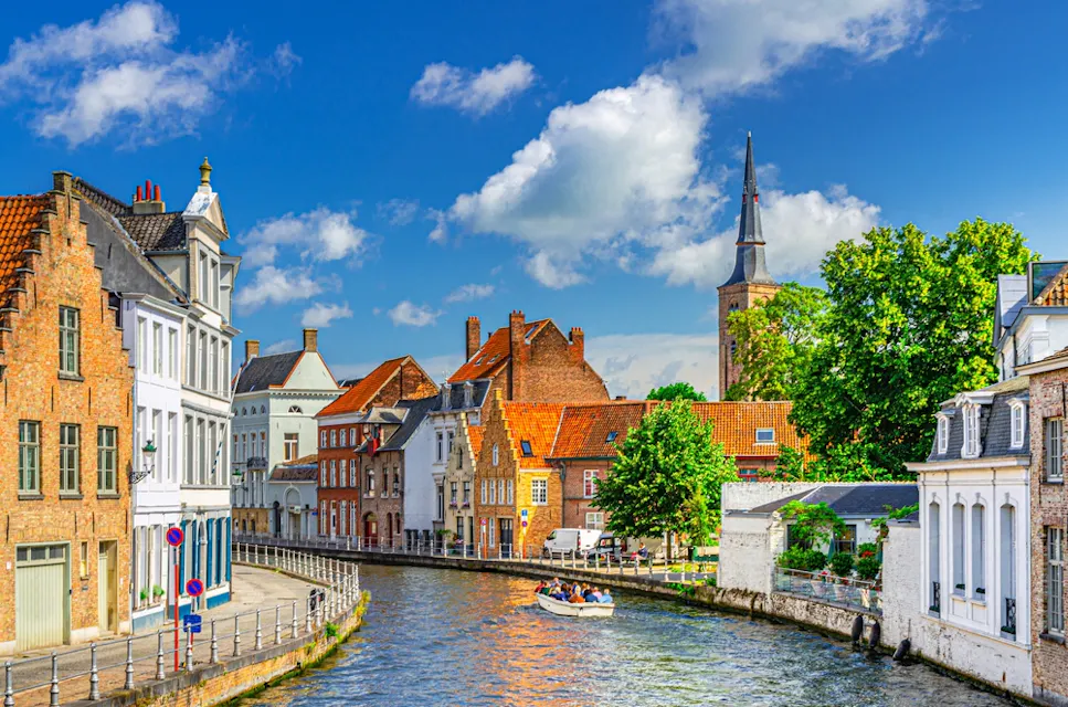 A picturesque canal in a European town with charming brick buildings and a church tower in the background. A small boat with people is cruising along the water under a partly cloudy blue sky. Lush greenery lines the canal.