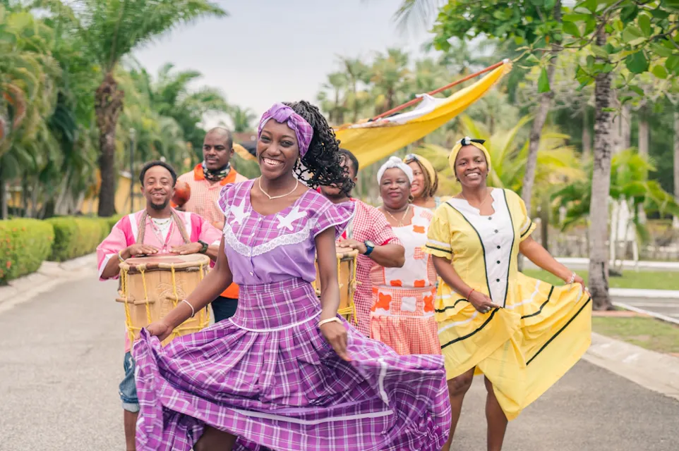 A group of people dressed in colorful, traditional clothing are joyfully dancing outdoors. They are surrounded by lush greenery and palm trees under a clear sky. A woman in a purple dress smiles while leading the group.