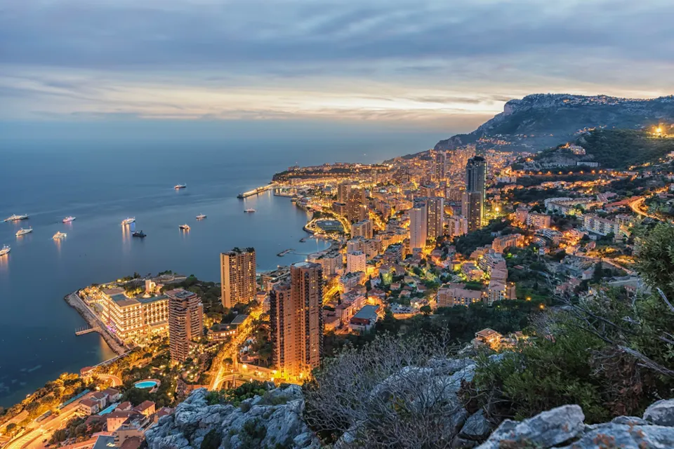 Aerial view of a coastal city at dusk, with buildings illuminated by lights. The sea is calm with boats dotting the waterfront. Hills surround the city, and the sky is cloudy with the last light of day.