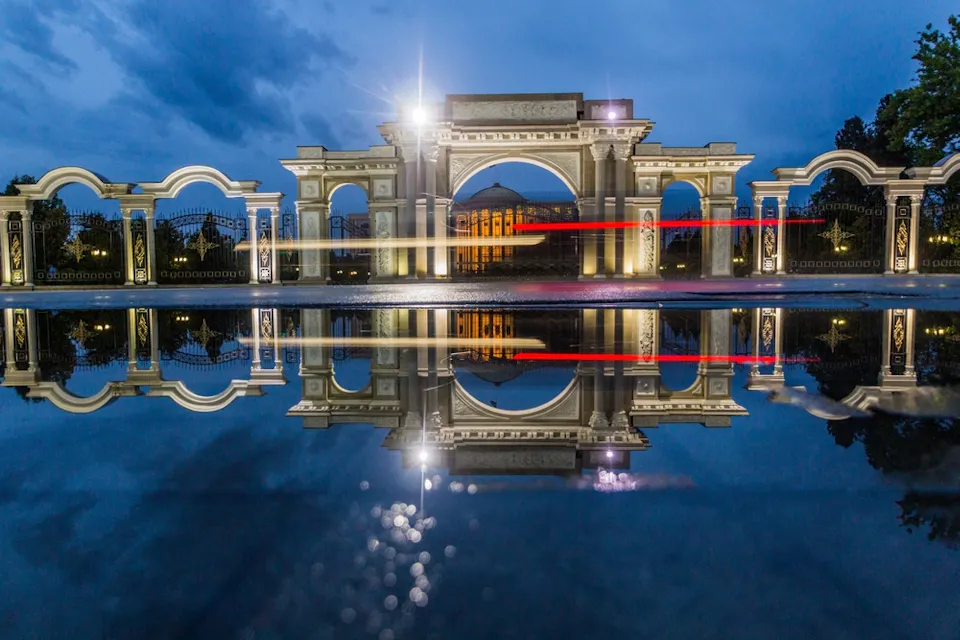 A grand architectural structure with multiple arches is beautifully lit at dusk, reflecting in a pool of water. The sky is a deep blue, and light trails from passing vehicles add a dynamic touch to the serene scene.