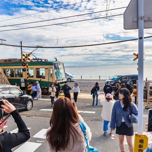 Getting from Tokyo to Kamakura People gather at a scenic railway crossing near a beach, taking photos as a train passes. The sky is partly cloudy, and the ocean is visible in the background. Some individuals hold smartphones to capture the moment.