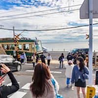 Getting from Tokyo to Kamakura People gather at a scenic railway crossing near a beach, taking photos as a train passes. The sky is partly cloudy, and the ocean is visible in the background. Some individuals hold smartphones to capture the moment.