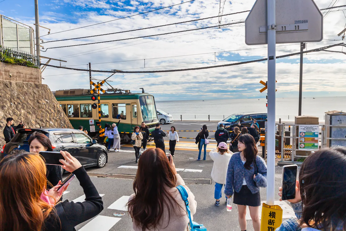 People gather at a scenic railway crossing near a beach, taking photos as a train passes. The sky is partly cloudy, and the ocean is visible in the background. Some individuals hold smartphones to capture the moment.
