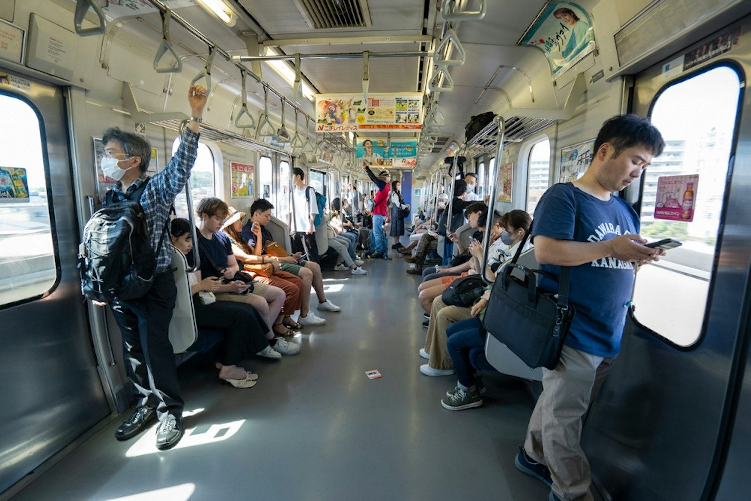 A crowded train interior with passengers seated and standing. Some people are using smartphones, while others gaze outside. The train is illuminated by natural light from large windows on both sides. Posters are visible above the seats.