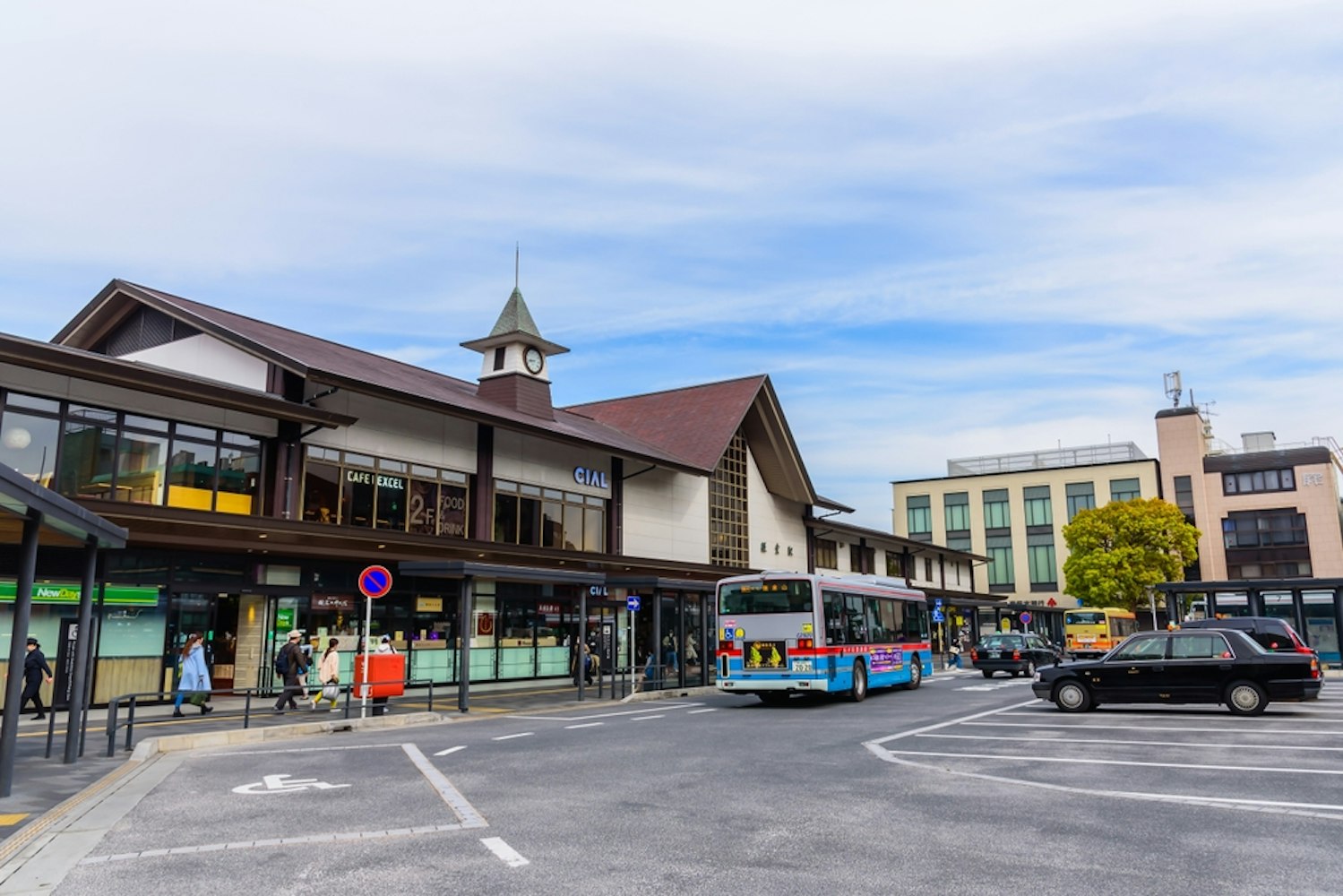 Outdoor view of a train station with a clock tower and multiple shops. Buses and taxis are parked in the foreground, while people walk nearby. The sky is partly cloudy, and modern buildings surround the area.