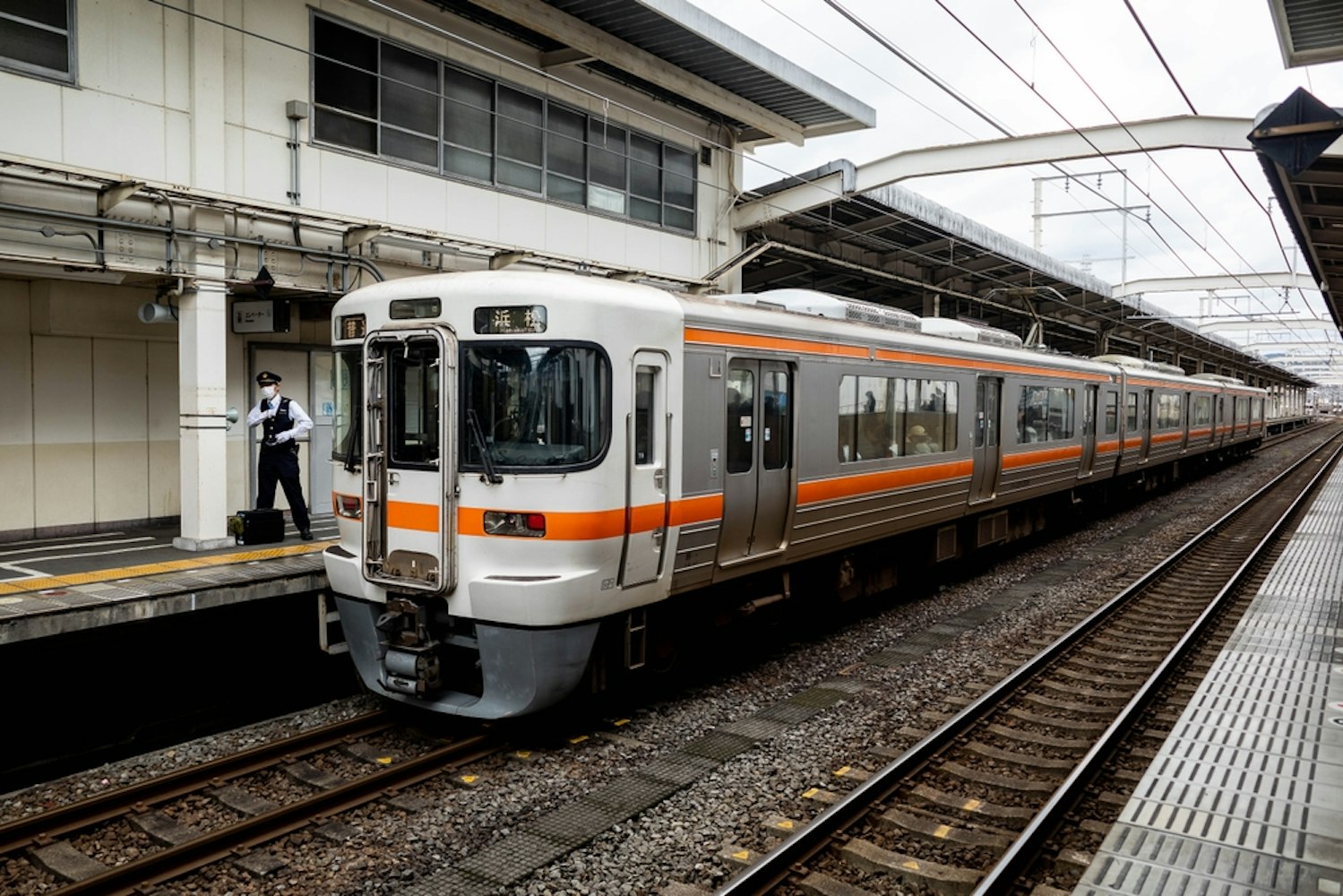 A passenger train with an orange stripe is parked at a platform in a station. A conductor checks the train doors. Overhead wires run above the tracks, and the station features a covered roof with support beams.
