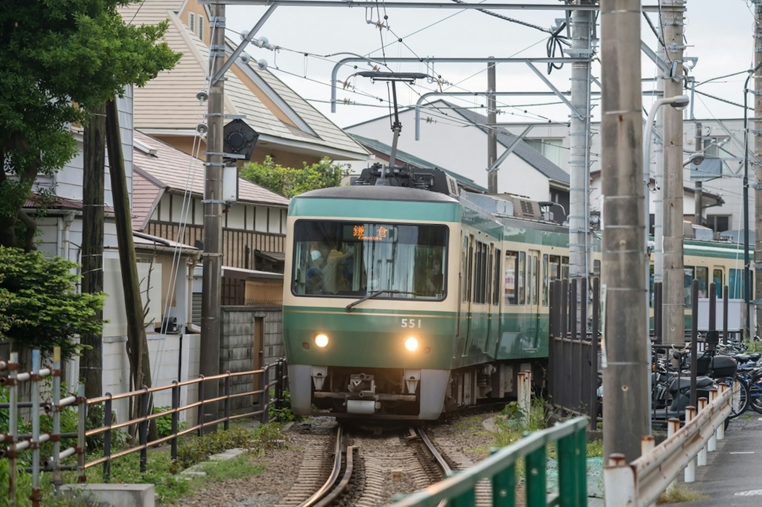 A green and cream-colored train travels through a narrow urban area, flanked by small houses and metal fences. Overhead power lines are visible, and a row of bicycles is parked alongside the track on the right.