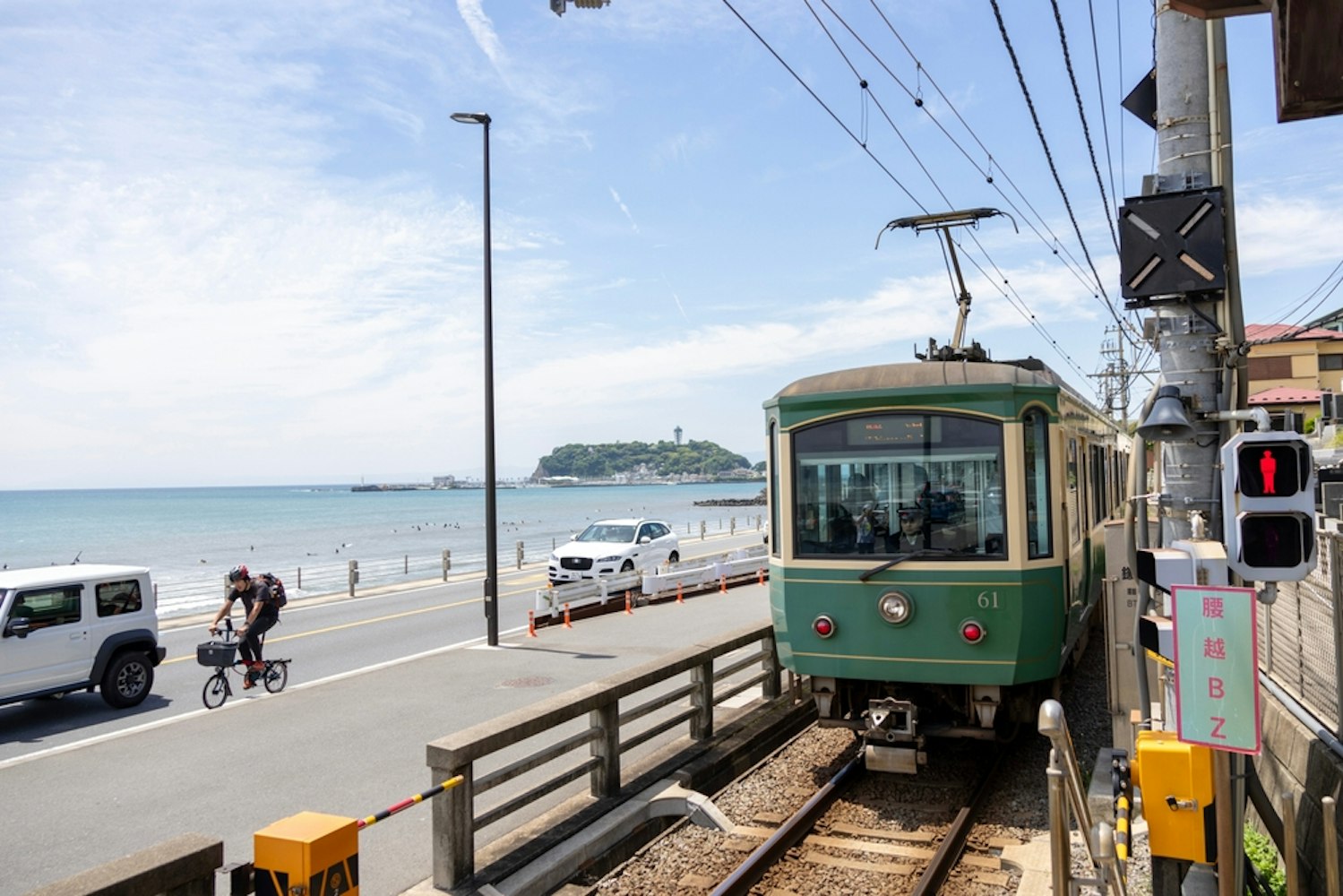 A green train travels along a seaside track with cars and a cyclist on the adjacent road. The ocean and a distant island with a tower are visible under a clear sky.