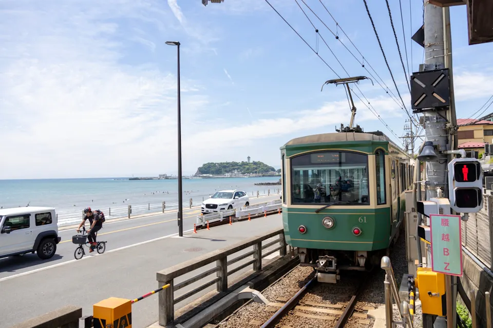 Exploring Kamakura by Train A green train travels along a seaside track with cars and a cyclist on the adjacent road. The ocean and a distant island with a tower are visible under a clear sky.