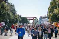 A lively crowd walks along a pathway lined with trees, leading to a large red torii gate in the background. Vendor stalls with colorful signs are on both sides, and the sky is clear and bright.