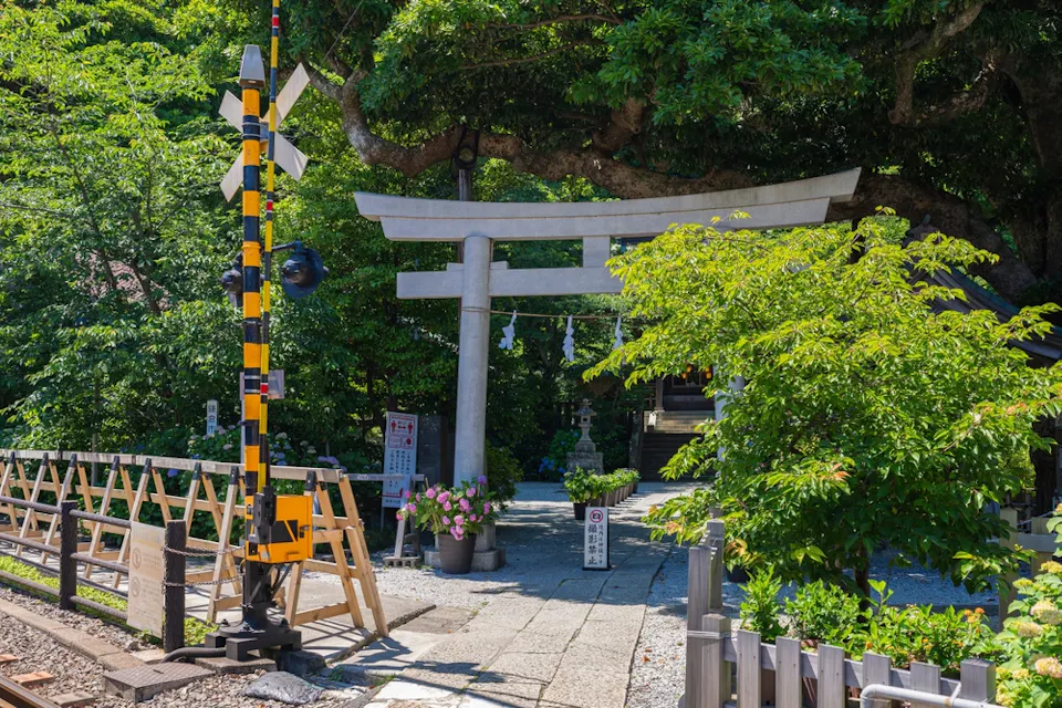 A Japanese shrine entrance with a torii gate situated near a railroad crossing. The scene is surrounded by lush green trees and vibrant flowers, with a clear blue sky overhead.