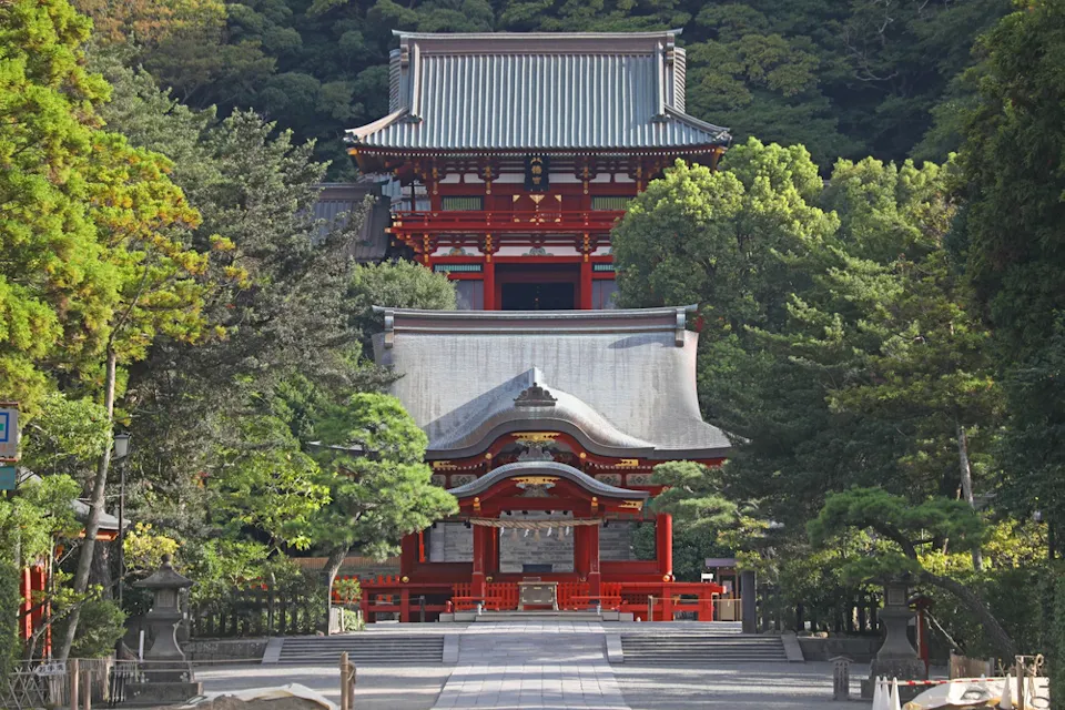 A traditional Japanese shrine surrounded by lush green trees, featuring intricate red and white wooden architecture with curved roofs. A wide stone path leads up to the entrance, framed by greenery.