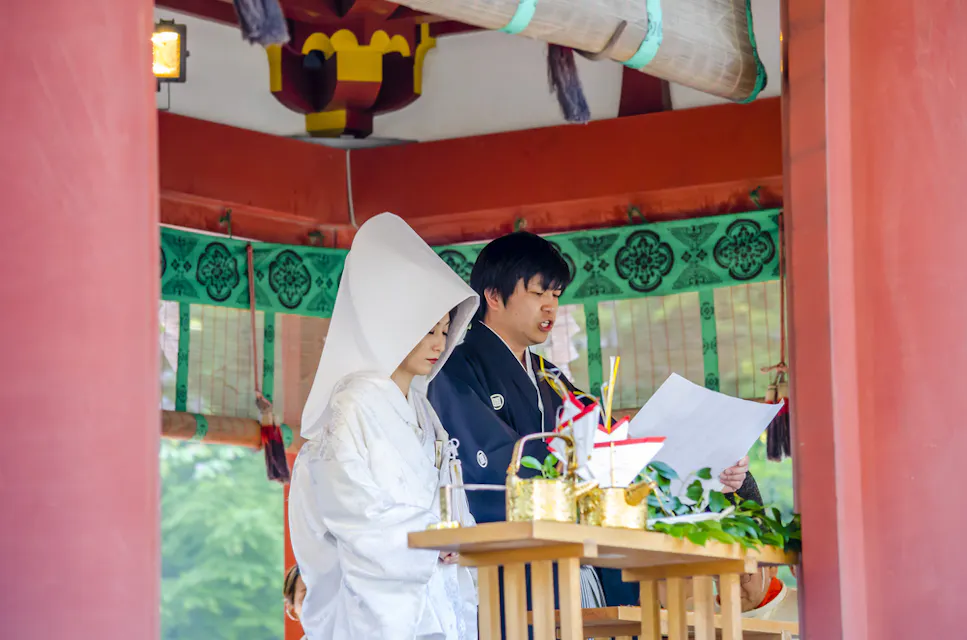 A couple in traditional Japanese wedding attire participate in a ceremony at a shrine. The bride wears a white kimono with a hood, and the groom wears a black kimono. They're standing at an altar adorned with decorations.