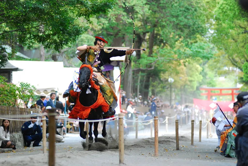 A person in traditional attire rides a galloping horse while shooting an arrow during a Yabusame archery event. Spectators watch from the sidelines, and lush green trees line the path.