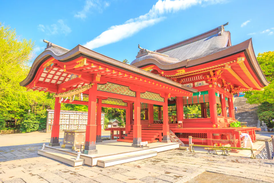 A beautiful traditional Japanese shrine with intricate red and white details stands against a clear blue sky. The wooden structure features ornate roof designs and is surrounded by lush greenery.