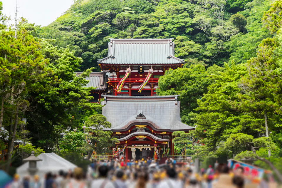 A vibrant scene of a traditional Japanese temple nestled among lush greenery. The temple roofs feature intricate designs, framed by dense trees, with a bustling crowd in the foreground.