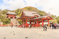 A traditional Japanese shrine with ornate red and white wooden structures is set against a backdrop of lush greenery. People are walking and exploring the temple grounds, and pigeons are on the foreground near the gravel path.