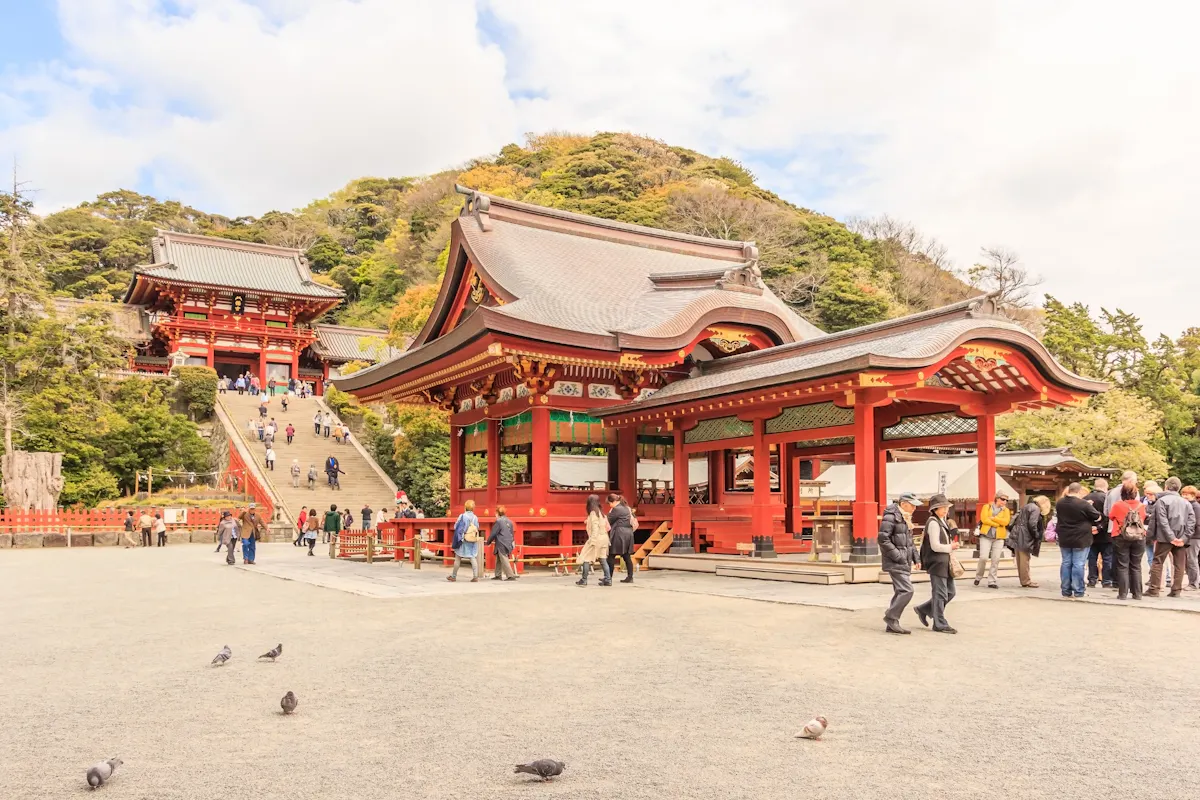 A traditional Japanese shrine with ornate red and white wooden structures is set against a backdrop of lush greenery. People are walking and exploring the temple grounds, and pigeons are on the foreground near the gravel path.