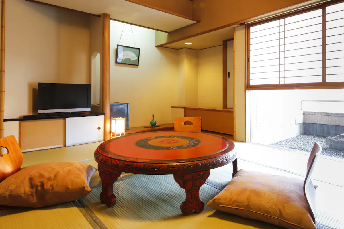Osaka Ryokan Kuramoto A traditional Japanese room features a round wooden table with intricate designs, surrounded by floor cushions on tatami mats. A television sits on a low shelf, and sliding doors open to a small outdoor area with gravel.