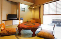 A traditional Japanese room features a round wooden table with intricate designs, surrounded by floor cushions on tatami mats. A television sits on a low shelf, and sliding doors open to a small outdoor area with gravel.