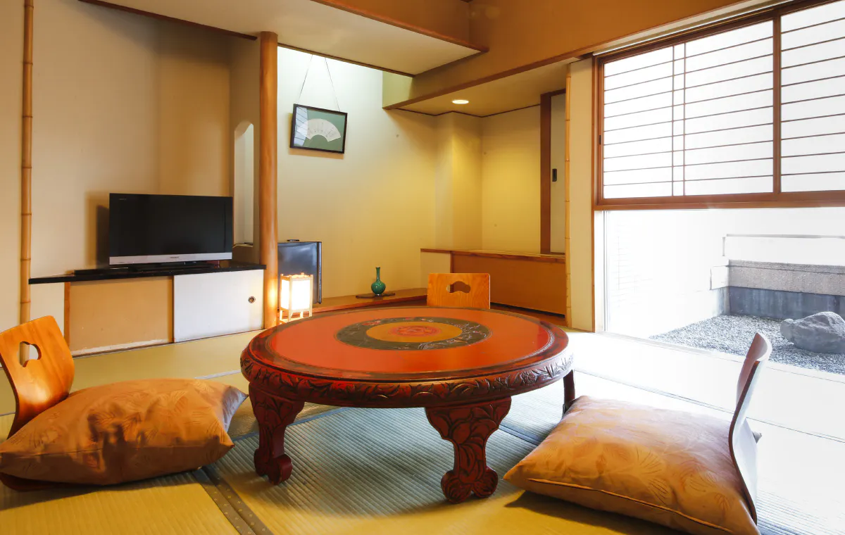 A traditional Japanese room features a round wooden table with intricate designs, surrounded by floor cushions on tatami mats. A television sits on a low shelf, and sliding doors open to a small outdoor area with gravel.