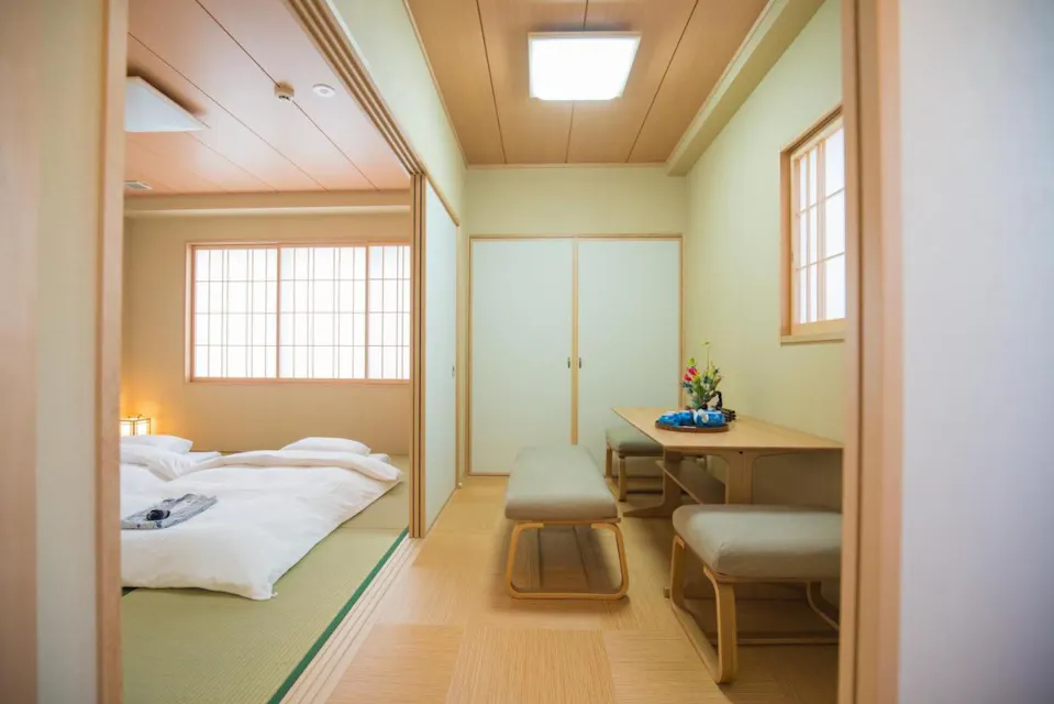 A traditional Japanese room with tatami mats, a low table, and cushions. To the left, there's a futon bed with a lamp nearby. The interior features sliding doors and soft lighting, creating a serene atmosphere.