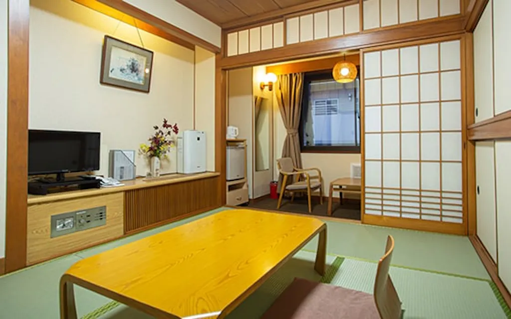 A cozy Japanese-style room with tatami mats and a low wooden table. Sliding shoji doors open to a small sitting area with two chairs and a window. The room features a TV, a vase with flowers, and warm lighting.