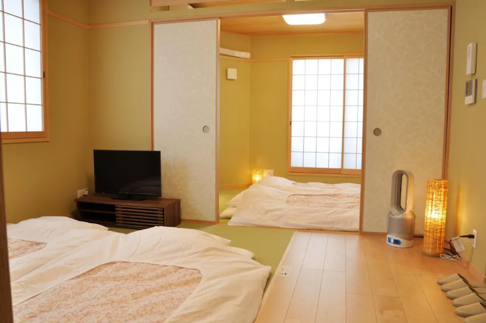 A traditional Japanese room with tatami flooring, futons, and sliding shoji doors. There are two neatly arranged futons on the floor, a small TV on a wooden stand, and a modern air purifier and lamp in the corner.