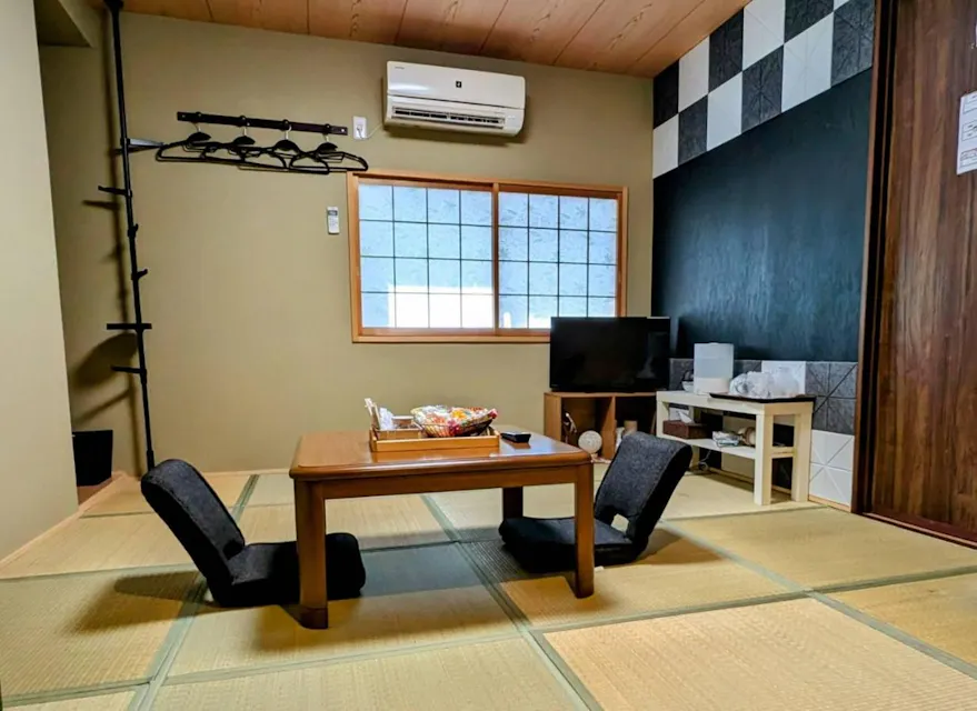 A traditional Japanese room with tatami flooring, a wooden table, and two floor chairs. A TV and shelves with items are against the wall. A window with paper panels, an air conditioner, and a decorative wall are visible.