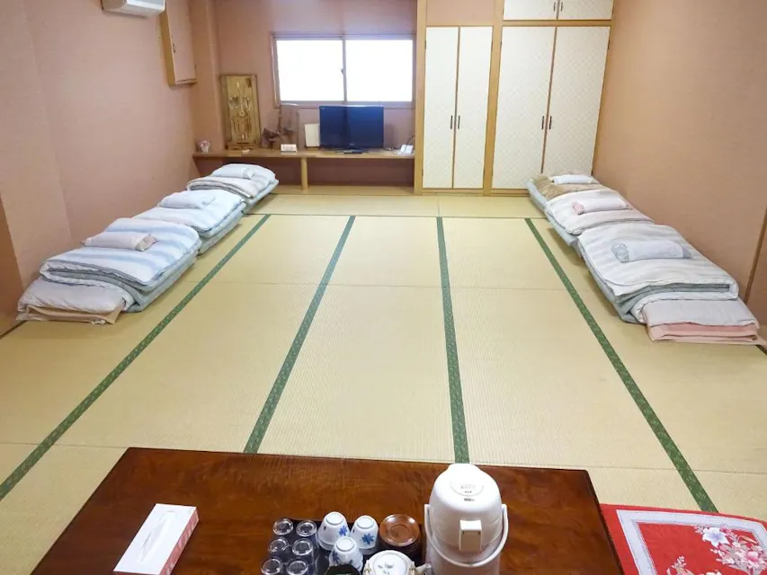 Tatami room with neatly arranged futons along two walls, a low wooden table in the center with cups and a kettle, and a small TV on a cabinet. The room has a soothing, traditional Japanese aesthetic.