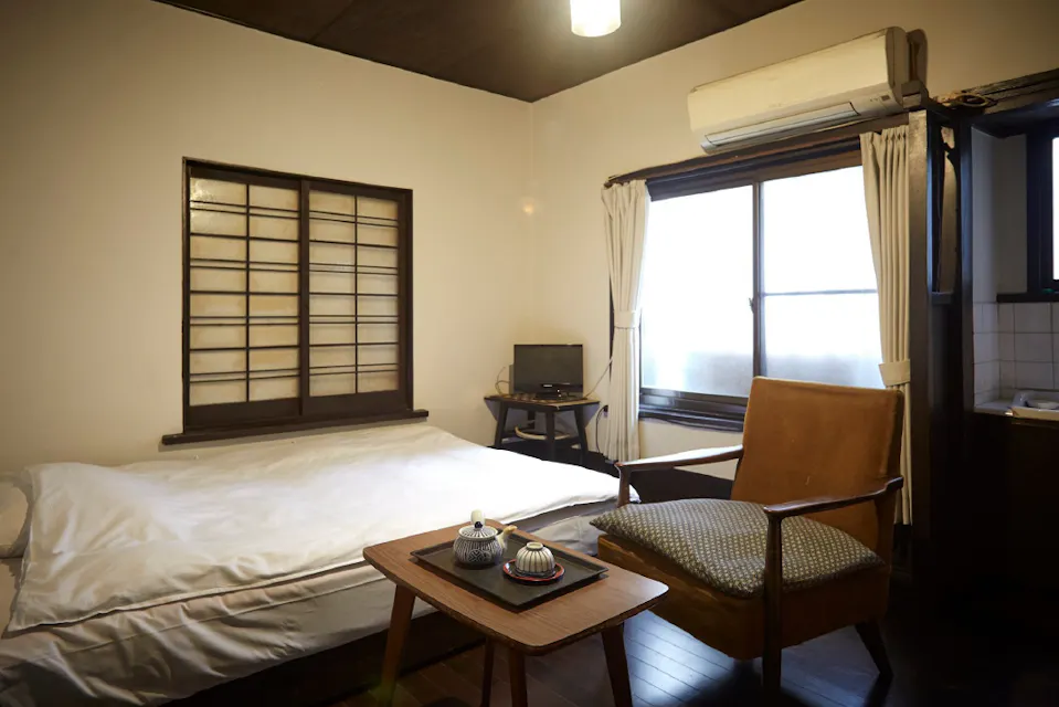A cozy room with a low bed, a wooden coffee table with tea set, and a cushioned chair. A small TV is by the window, which has white curtains. The room features wooden flooring, a shoji-style window, and an air conditioner.