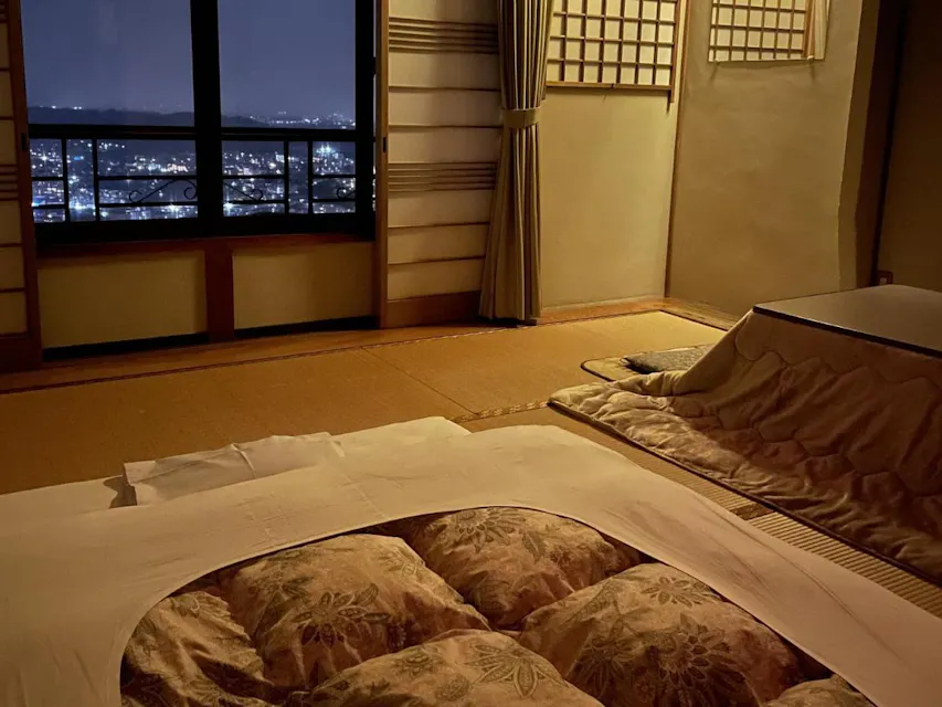A cozy room with tatami mats, a futon bed, and a kotatsu table. Large windows reveal a night cityscape. Traditional Japanese elements like shoji screens and wooden framework are visible. The curtain is partially drawn.