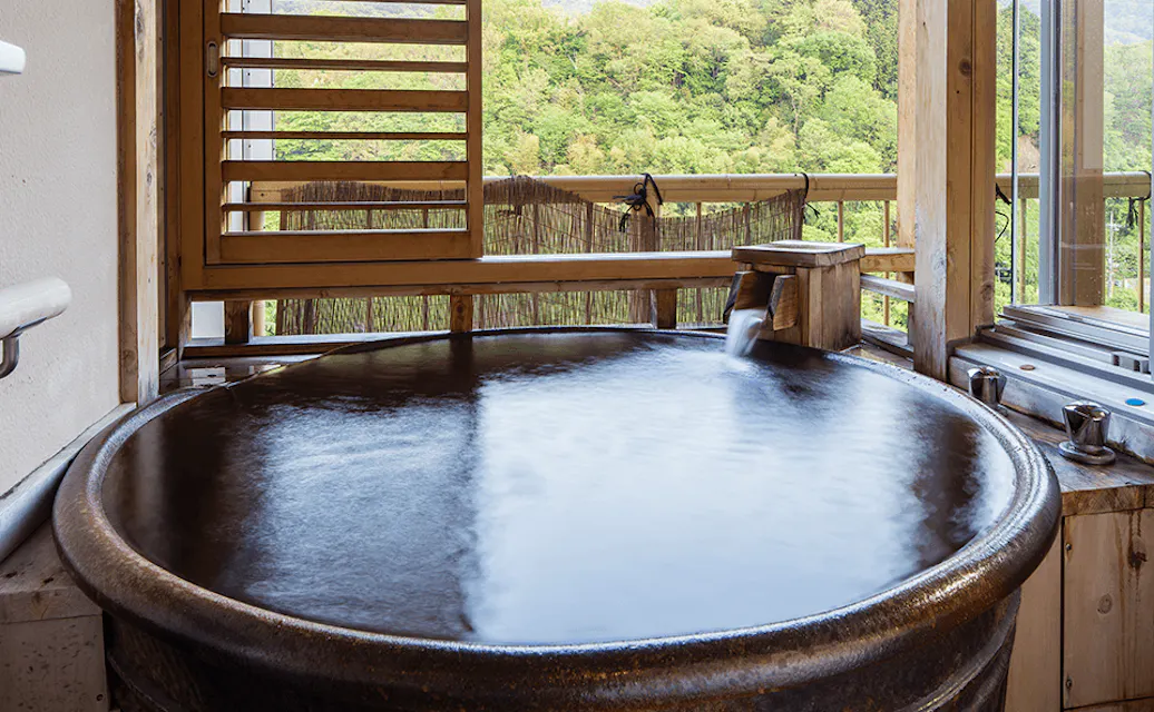 A round, steaming hot tub fills with water from a wooden spout in a cozy room. Large windows frame a lush green forest, offering a serene, natural view. Wooden shutters partially cover the windows, creating a tranquil, inviting atmosphere.