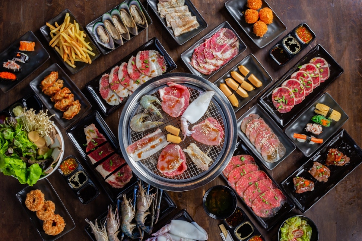 Shabu and Grill traditional japanese food A top view of a variety of Korean BBQ dishes on a wooden table. The spread includes raw meats, seafood, vegetables, dumplings, sauces, and garnishes arranged around a grill, with side dishes and condiments in black plates.