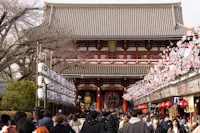 A bustling crowd walks towards a traditional Japanese temple, with lanterns and cherry blossom decorations lining the path. The temple's grand facade features intricate designs, and bare trees are visible on the side.