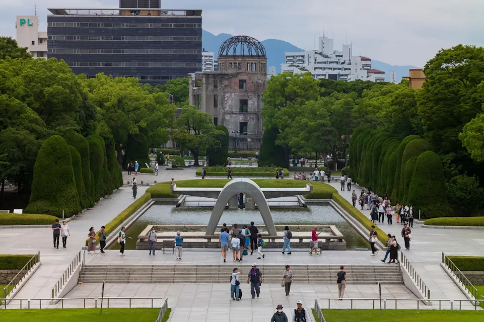 People walk through Hiroshima Peace Memorial Park with the Genbaku Dome in the background. The park is lush with green trees and features a notable arch monument over a reflective pool, on a cloudy day.