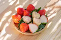 A wooden plate on a light wooden table holds an arrangement of ripe red and pale white strawberries, some of which are halved, with leaves still attached. Dappled sunlight creates patterns of shadows on the surface.