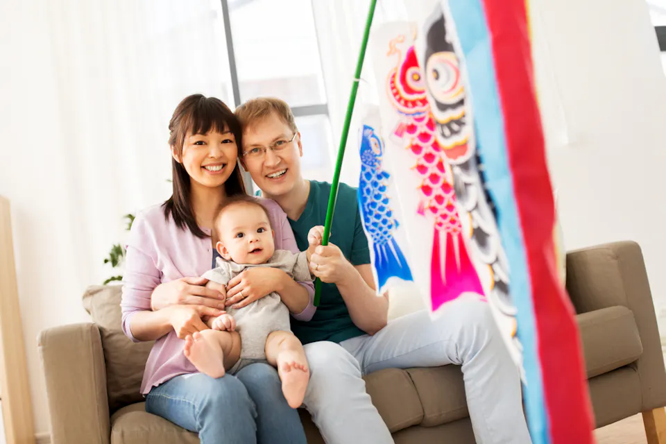 A smiling family of three sits on a couch. The mother holds a baby on her lap, while the father holds a colorful carp streamer. The room is bright and airy, with a large window in the background.