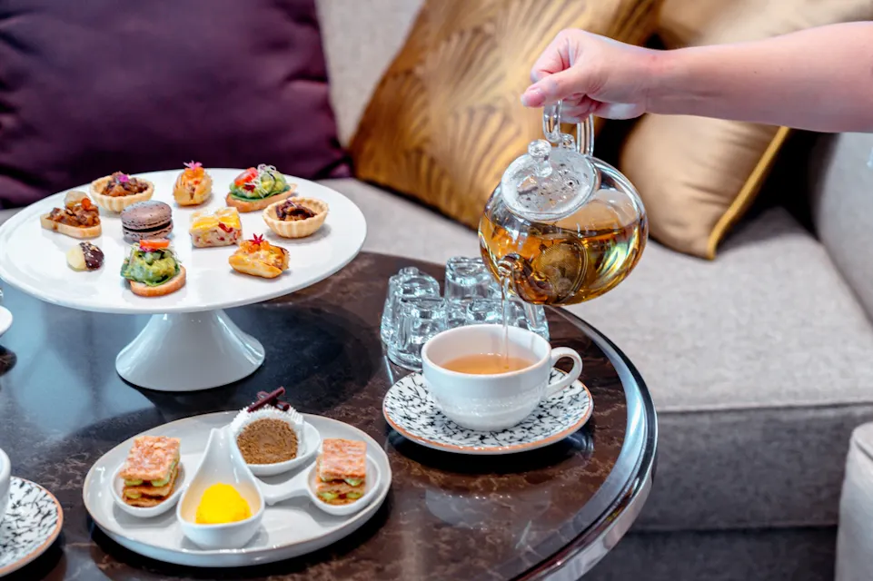 A person pours tea from a glass teapot into a decorated teacup. The table is set with plates of assorted desserts and sandwiches on elegant stands. The setting includes comfortable seating with pillows in the background.