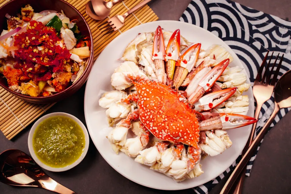 A plate of steamed crab with orange shell and white meat is served alongside a bowl of spicy salad, a dish of green sauce, and utensils on a patterned tablecloth.