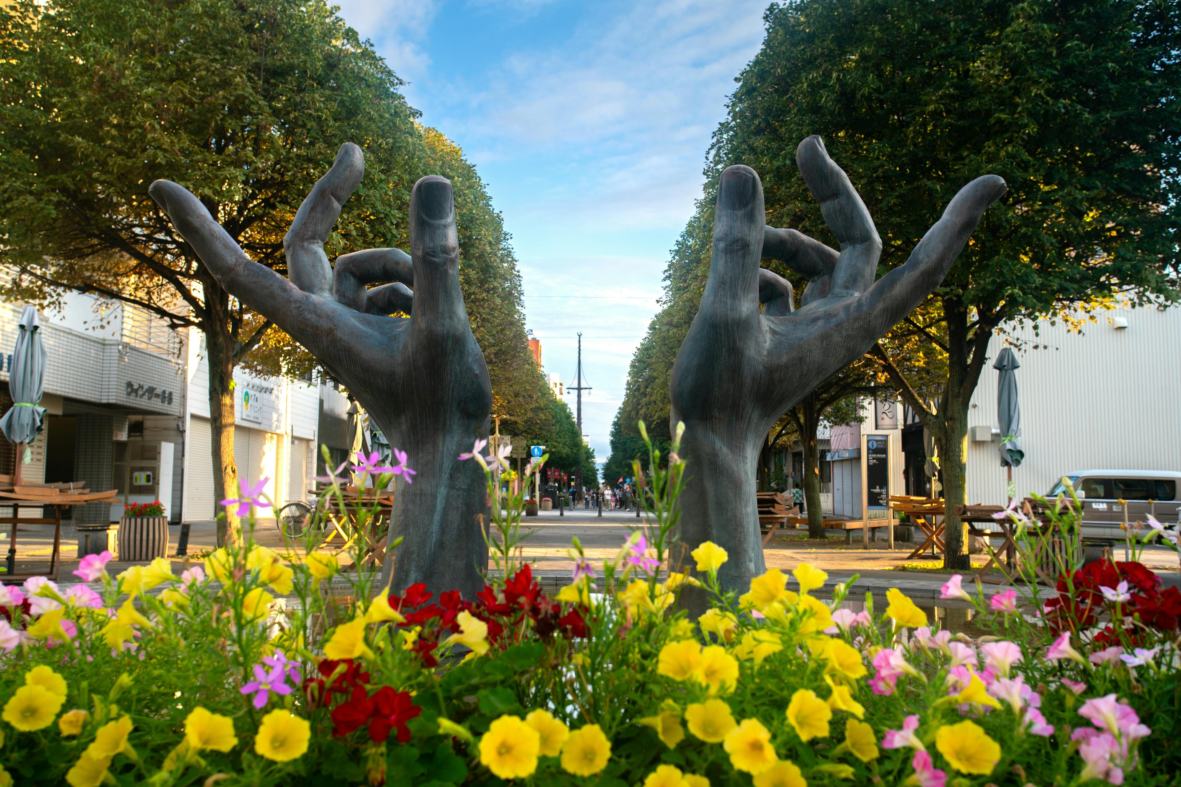 A large sculpture of two hands making the "OK" gesture stands in the middle of a street, surrounded by colorful flowers and trees under a blue sky. Tables and chairs line the sidewalks.