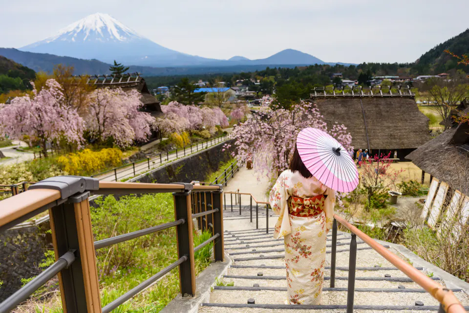 A person wearing a floral kimono walks down stairs holding a pink parasol, with cherry blossoms, traditional thatched-roof houses, and a snow-capped mountain in the background, under a cloudy sky.