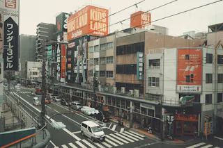 A busy urban street in Japan with cars, pedestrians, and tall buildings covered in colorful signs and advertisements in Japanese. Shops line the street, and a crosswalk is visible in the foreground.