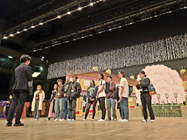 A group of people stand in a line on a stage, facing a person holding a sheet of paper. Behind them is a colorful set with a cherry blossom tree and illuminated backdrop. The setting appears to be a theater or auditorium.