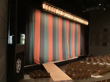 A theater stage with a closed, vertically striped curtain in teal and orange. Rows of empty wooden seats face the stage, and a walkway extends into the audience. Overhead lights illuminate the curtain.