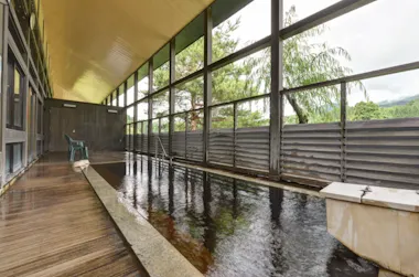 A long, narrow indoor hot spring bath with large windows, wooden floors, and outdoor views of trees and mountains. A plastic chair sits near the far end.