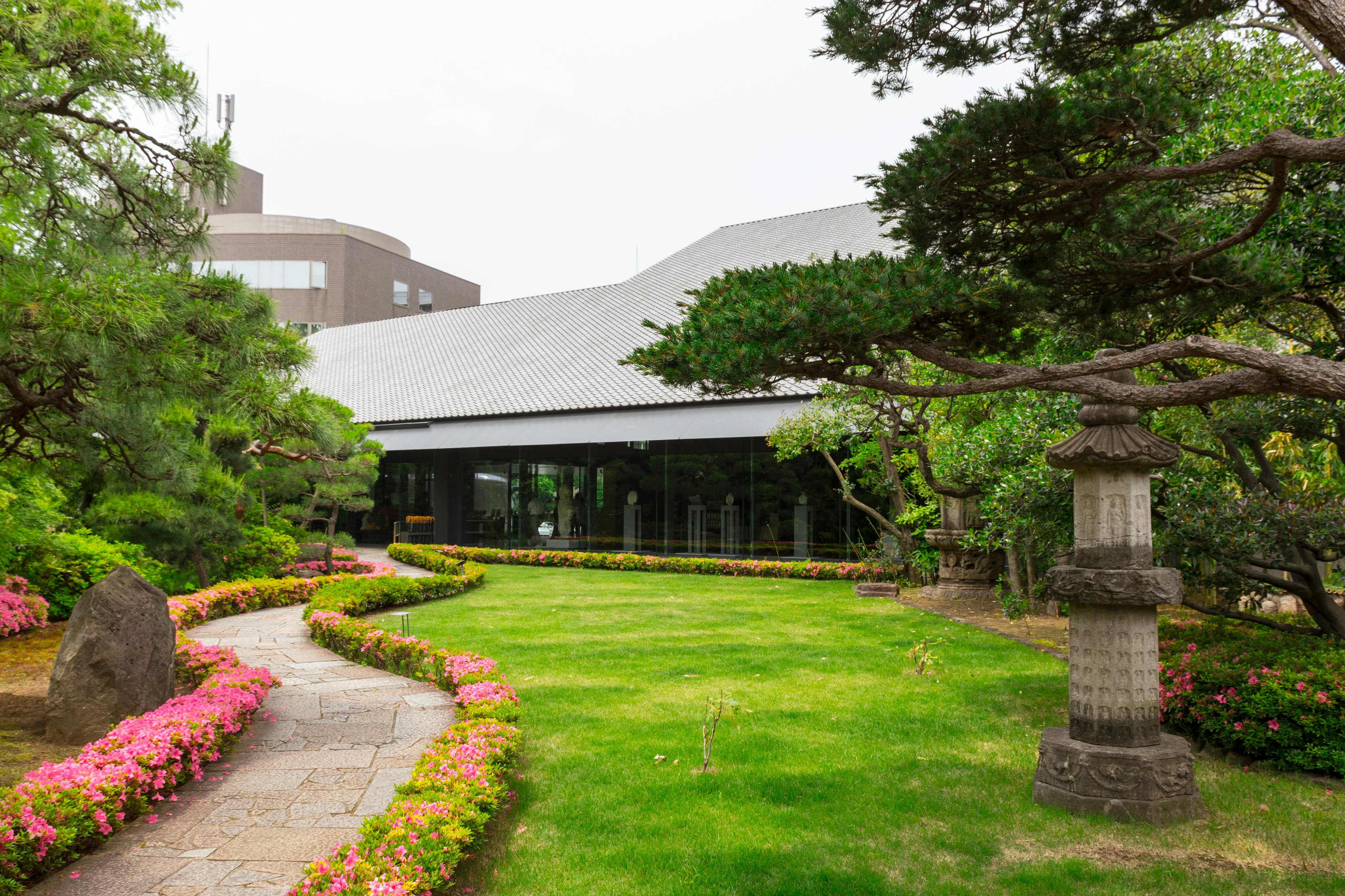 A stone path lined with pink flowers winds through a manicured Japanese garden with green grass, trees, and a traditional stone lantern near a modern building.