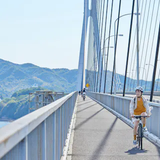 Shimanami Kaido (Nishiseto Expressway) A person wearing a helmet rides a bicycle on a bridge with mountains and water visible in the background; other cyclists are seen further along the bridge under a clear blue sky.